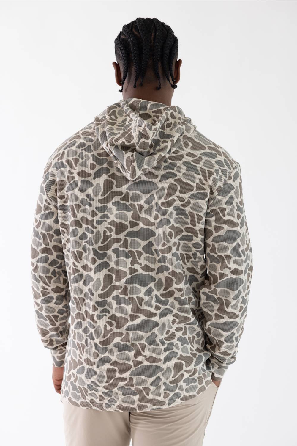 A man viewed from behind wearing a long-sleeve hooded sweatshirt with a camouflage pattern in beige, gray, and brown, paired with light beige pants against a plain white studio background.