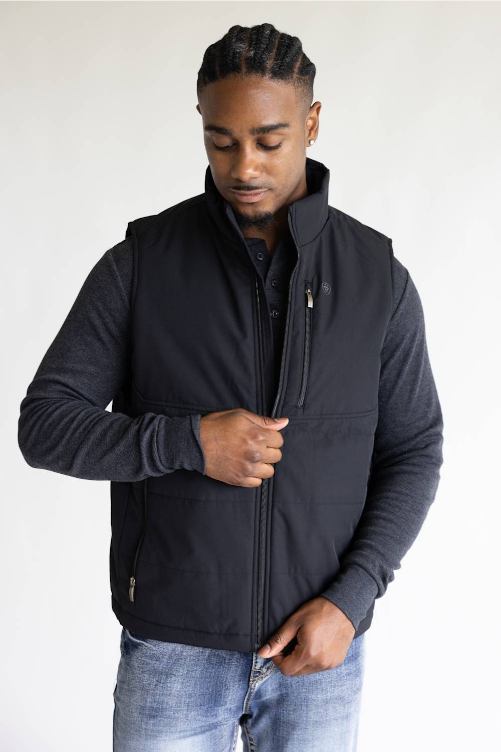 A man wearing a black zip-up fleece vest over a dark gray long-sleeve shirt and blue jeans, standing against a plain light background.