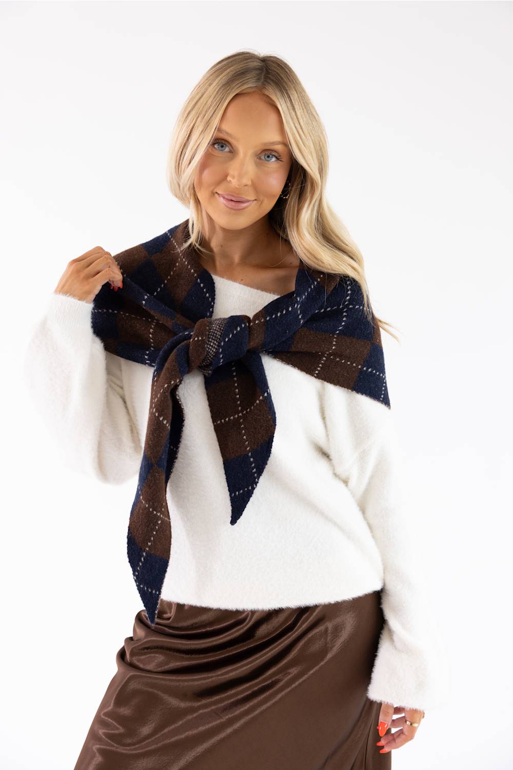 A blonde woman wearing a white knit sweater and a brown satin skirt, modeling a dark brown and navy plaid scarf tied around her neck against a plain white background.