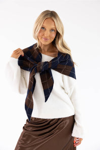 A blonde woman wearing a white knit sweater and a brown satin skirt, modeling a dark brown and navy plaid scarf tied around her neck against a plain white background.
