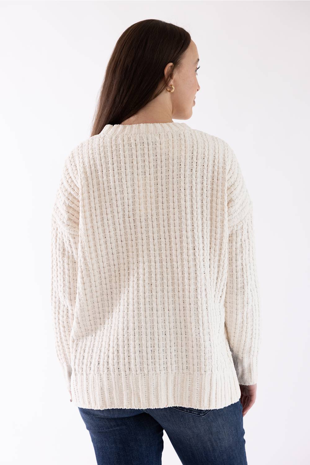A woman viewed from the back wearing a cream-colored, textured knit sweater and blue jeans against a plain white background.