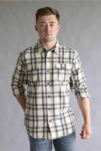 Front three-quarter view of a male model with one hand behind his back wearing the Split Flannel Shirt for Men in Grey Ecru, showcasing the beige, gray, and brown plaid pattern, long sleeves, and dual chest button pockets against a neutral gray studio background.