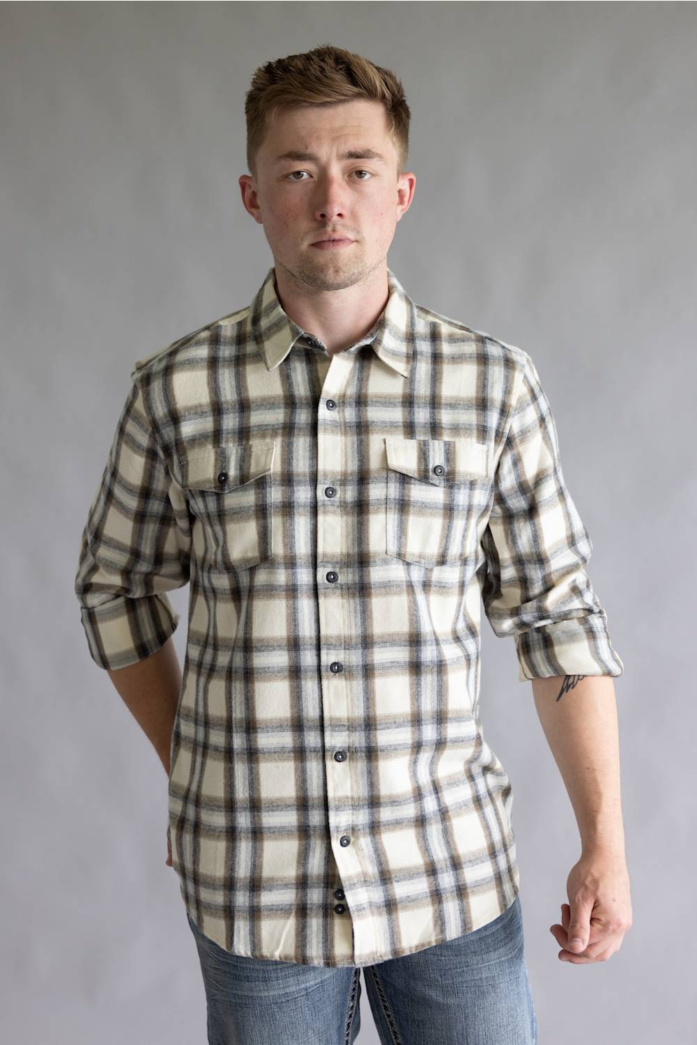 Front three-quarter view of a male model with one hand behind his back wearing the Split Flannel Shirt for Men in Grey Ecru, showcasing the beige, gray, and brown plaid pattern, long sleeves, and dual chest button pockets against a neutral gray studio background.