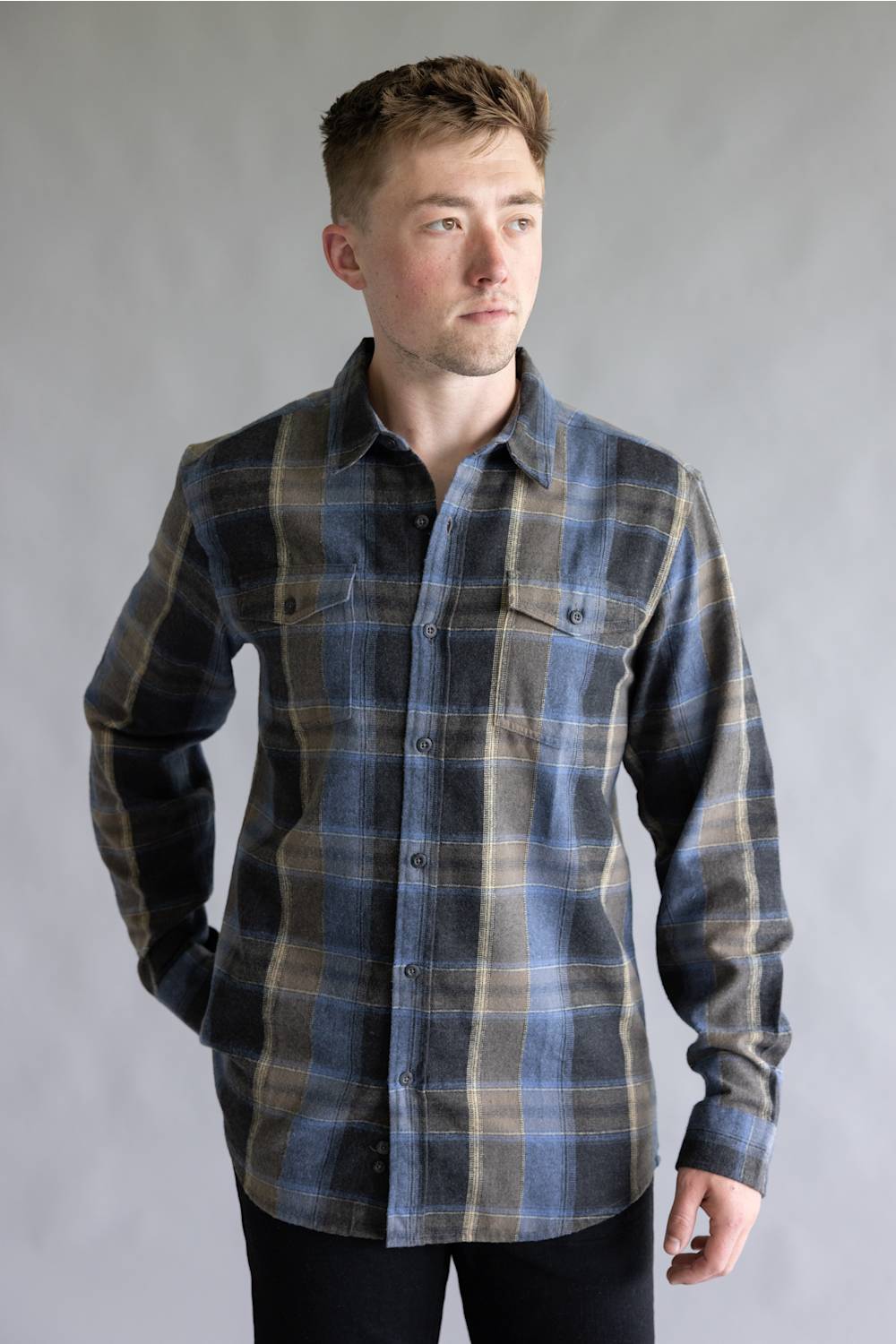 Close-up front view of a male model with hand in pocket wearing the Split Flannel Shirt for Men in Moka, showcasing the blue, dark gray, and tan plaid pattern, button-down collar, and dual chest pockets against a neutral gray background.