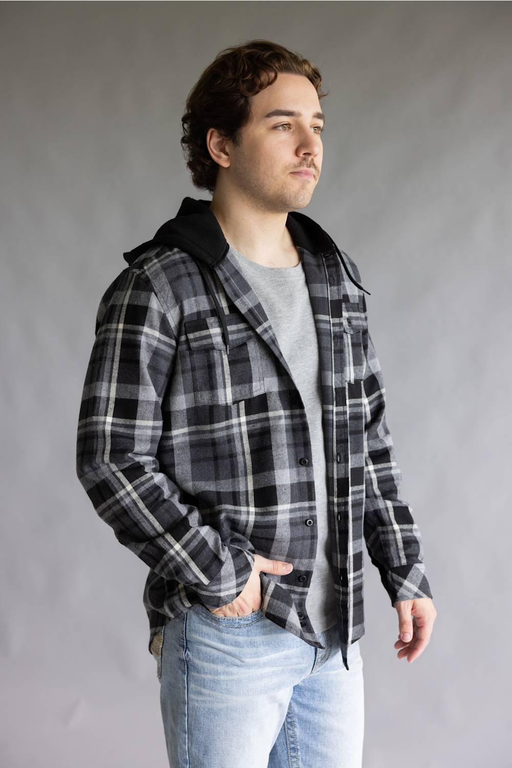 Model posing at a three-quarter angle wearing an open, unbuttoned Split Men's long-sleeve hooded flannel shirt in a black, grey, and white plaid pattern, styled over a grey t-shirt with light wash denim jeans against a grey studio background.