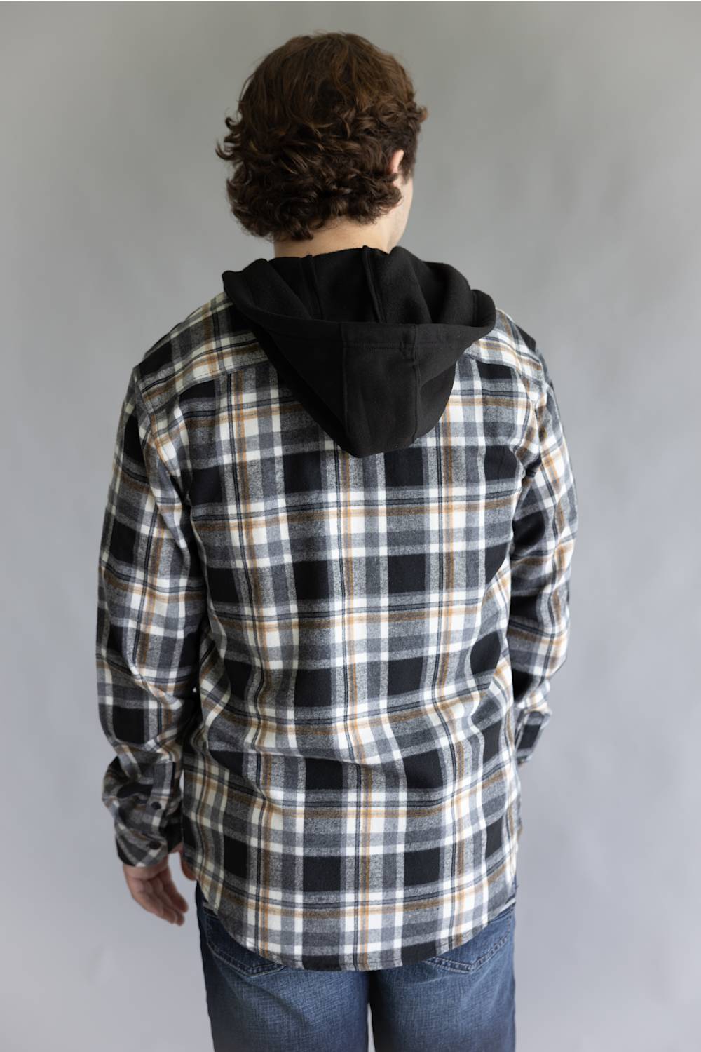 Back view of a model wearing the Split Men's long-sleeve hooded flannel shirt in a cream, black, and tan plaid pattern, featuring an attached solid black hood and straight back yoke against a gray studio background.