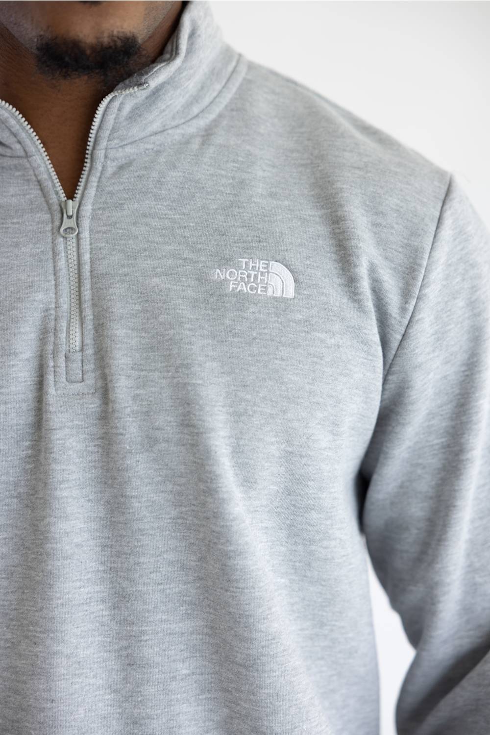 A close up of a man wearing a heather grey quarter zip The North Face men's sweatshirt with a white logo on the upper left chest.