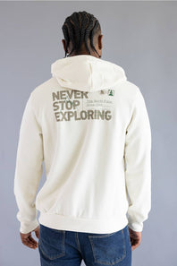 A man viewed from behind wearing an off-white hoodie with 'NEVER STOP EXPLORING' printed in brown across the back, paired with blue jeans against a gray studio background.