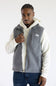 A man wearing a gray fleece vest by The North Face over a cream hoodie and blue jeans, standing against a plain white studio background.