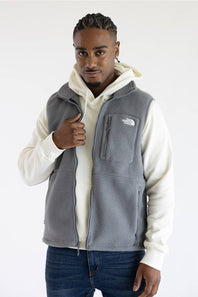 A man wearing a gray fleece vest by The North Face over a cream hoodie and blue jeans, standing against a plain white studio background.