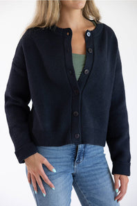 Front ¾ view of a female model wearing the Things Between Alyssa Cardigan in Navy, featuring a loose fit, large dark buttons, dropped shoulders, and ribbed hem, styled over a sage green top and light-wash blue jeans.