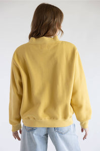 A person shown from the back wearing a mustard yellow crewneck sweatshirt and light blue jeans against a plain light gray studio background.