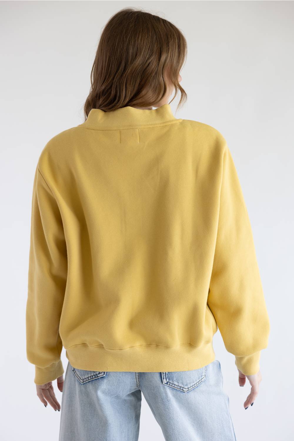 A person shown from the back wearing a mustard yellow crewneck sweatshirt and light blue jeans against a plain light gray studio background.