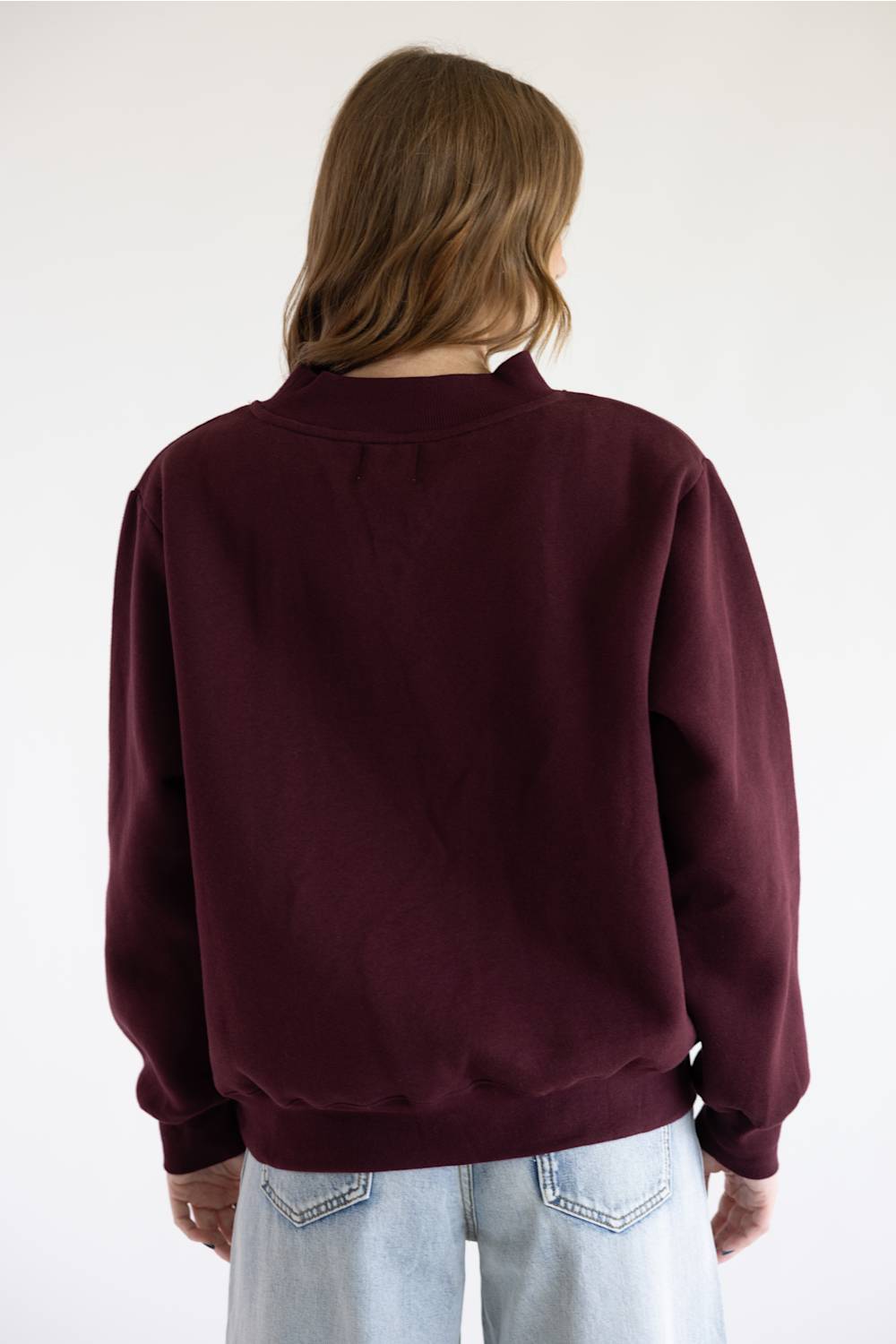 A close up of a woman with red hair wearing a loose-fitting, long-sleeve crewneck sweater in deep burgundy and blue jeans, standing against a plain white background.