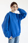 A woman wearing an oversized royal blue sweatshirt and light blue jeans, posing against a plain white studio background.