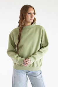 A woman with long light brown hair in a side braid wears a sage green crewneck sweatshirt and light wash blue jeans, standing against a plain white studio background.