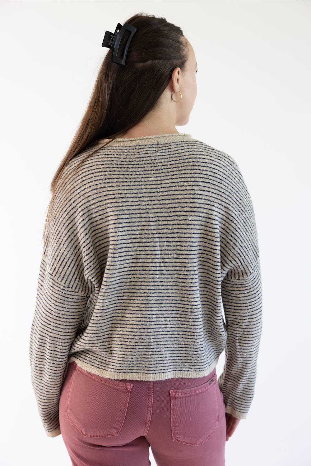 A woman viewed from the back wearing a beige  knit sweater with horizontal stripes and dusty rose pants against a plain white background.