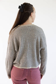 A woman viewed from the back wearing a beige  knit sweater with horizontal stripes and dusty rose pants against a plain white background.