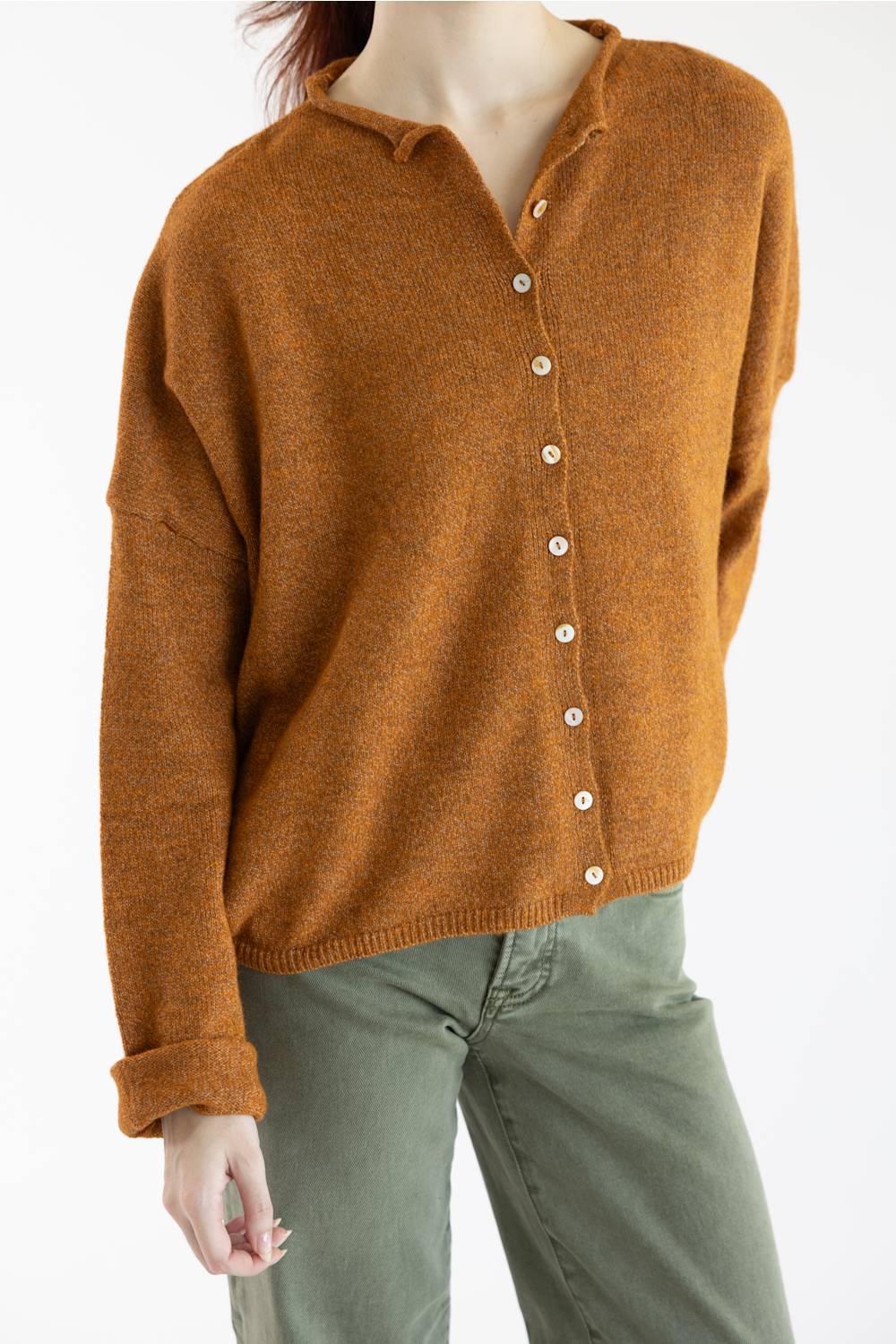 A woman wearing a long-sleeve knit cardigan in a warm rust-orange color with white buttons, paired with olive green pants, against a plain white background.