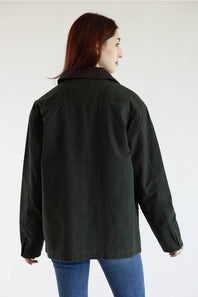 A woman with red hair is shown from the back wearing a dark olive green utility jacket with a brown collar, paired with blue jeans, against a plain white studio background.