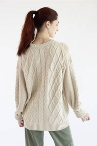 A woman wearing a cream-colored cable-knit sweater and olive green pants, shown from the neck down against a plain white background.