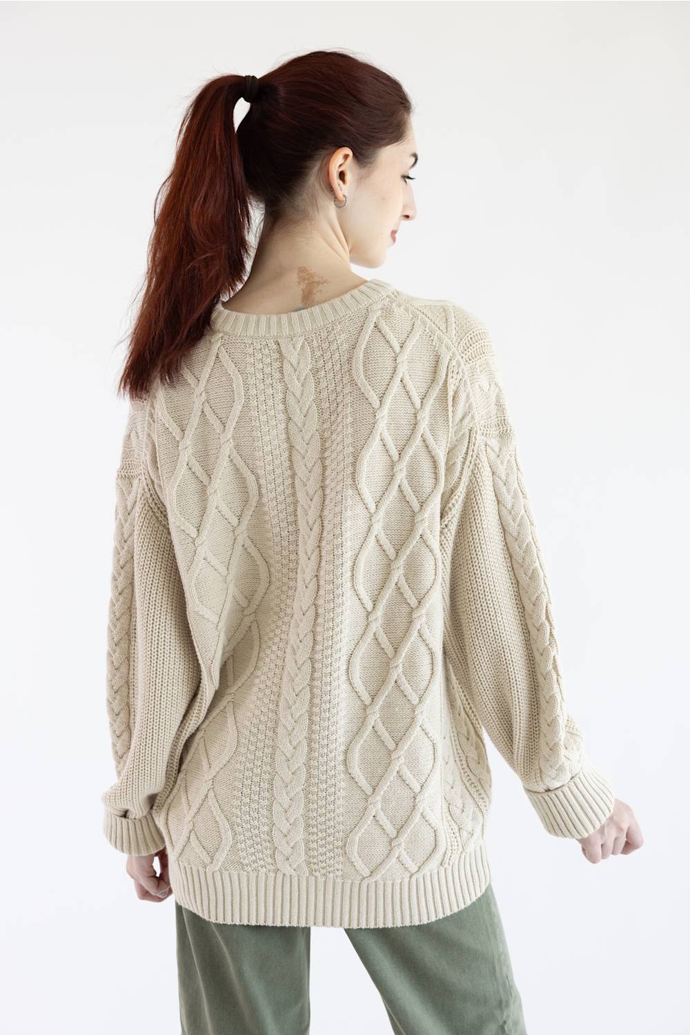A woman wearing a cream-colored cable-knit sweater and olive green pants, shown from the neck down against a plain white background.