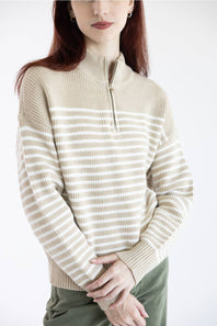 A woman wearing a beige and off-white horizontally striped quarter-zip knit sweater, paired with olive green pants, against a plain white studio background.