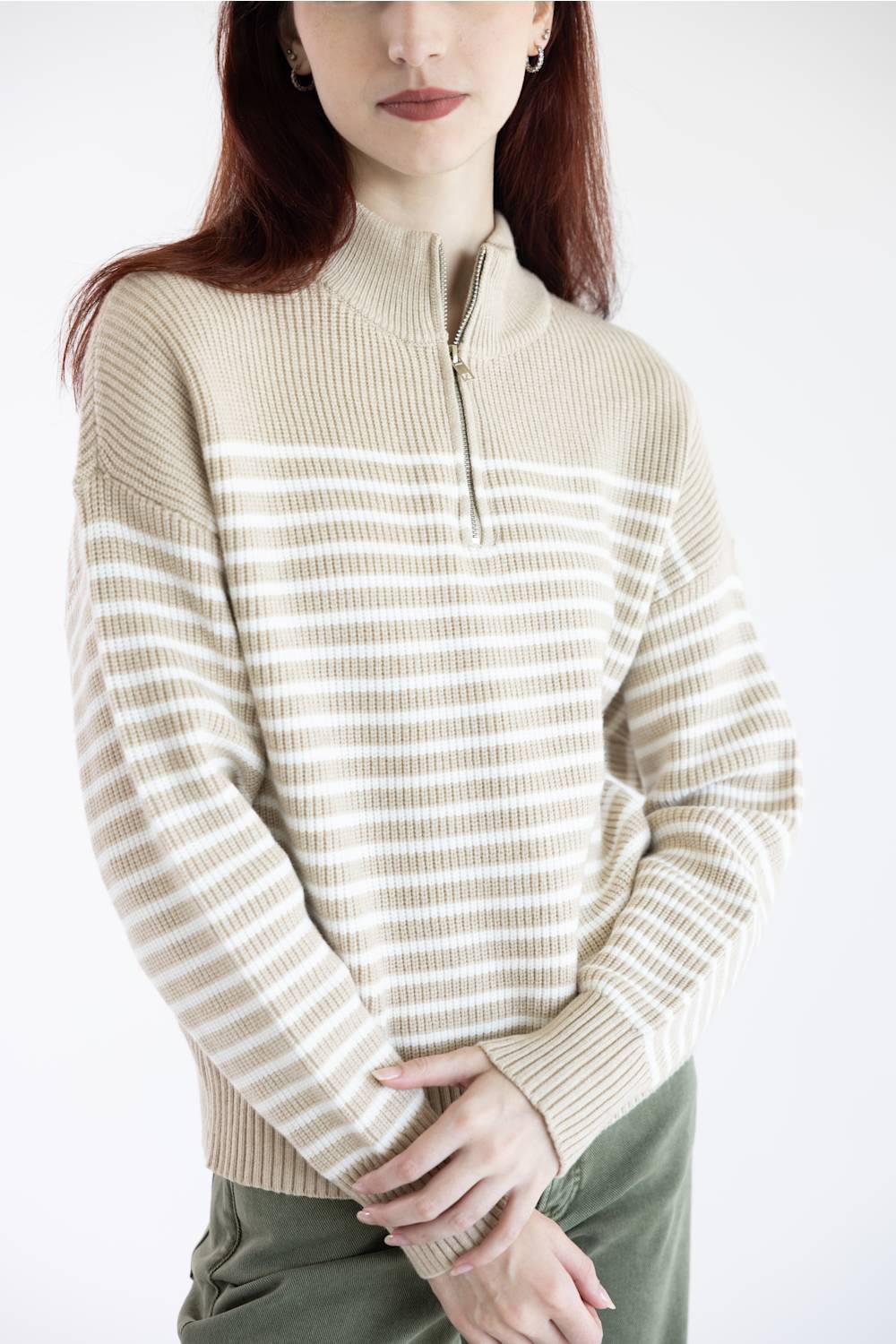 A woman wearing a beige and off-white horizontally striped quarter-zip knit sweater, paired with olive green pants, against a plain white studio background.