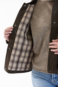 A woman modeling a dark brown suede jacket with a dark brown corduroy collar, worn over a brown knit sweater and blue jeans, against a plain white studio background.