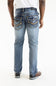 Rear view of a person wearing light-wash blue denim jeans with decorative gold-toned stitching and pocket detailing, paired with a white shirt and brown shoes, against a plain white studio background.