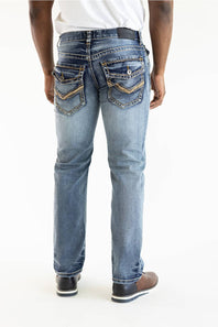 Rear view of a person wearing light-wash blue denim jeans with decorative gold-toned stitching and pocket detailing, paired with a white shirt and brown shoes, against a plain white studio background.