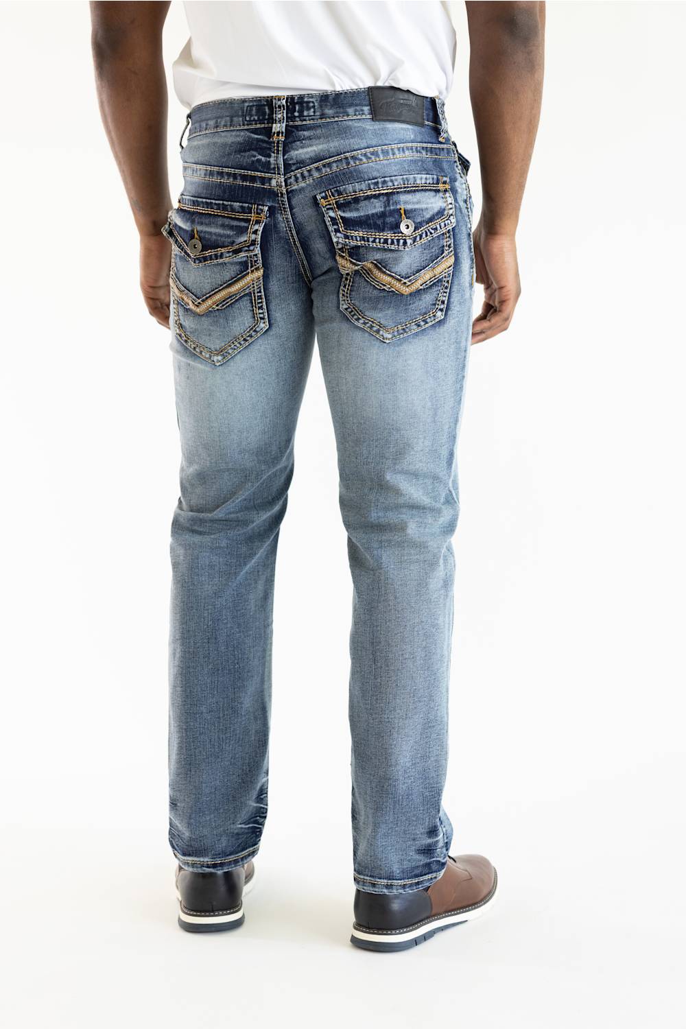 Rear view of a person wearing light-wash blue denim jeans with decorative gold-toned stitching and pocket detailing, paired with a white shirt and brown shoes, against a plain white studio background.