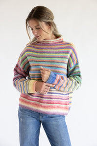 Woman wearing a long sleeve striped multi rainbow colored sweater with blue jeans standing in front of a white background