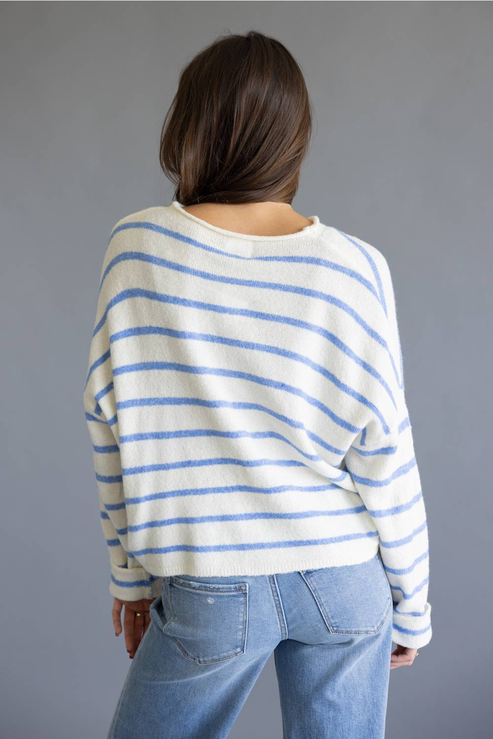 A woman viewed from behind wearing a cream-colored knit sweater with thin blue horizontal stripes and light-wash blue jeans, set against a solid gray studio background.