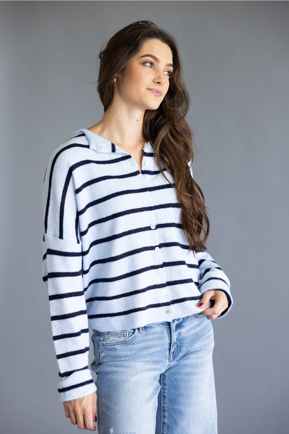 A woman wearing a white long-sleeve knit top with navy blue horizontal stripes, paired with light blue jeans, posing against a solid gray studio background.
