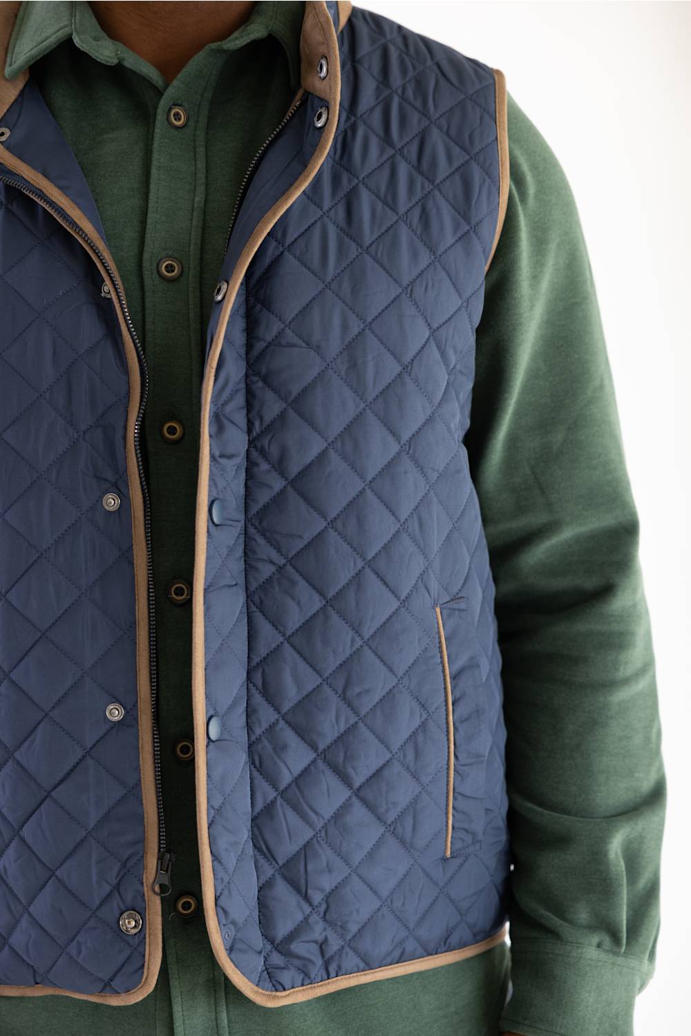 A close-up of a person wearing a navy blue diamond-quilted vest with tan trim over a green button-up shirt.