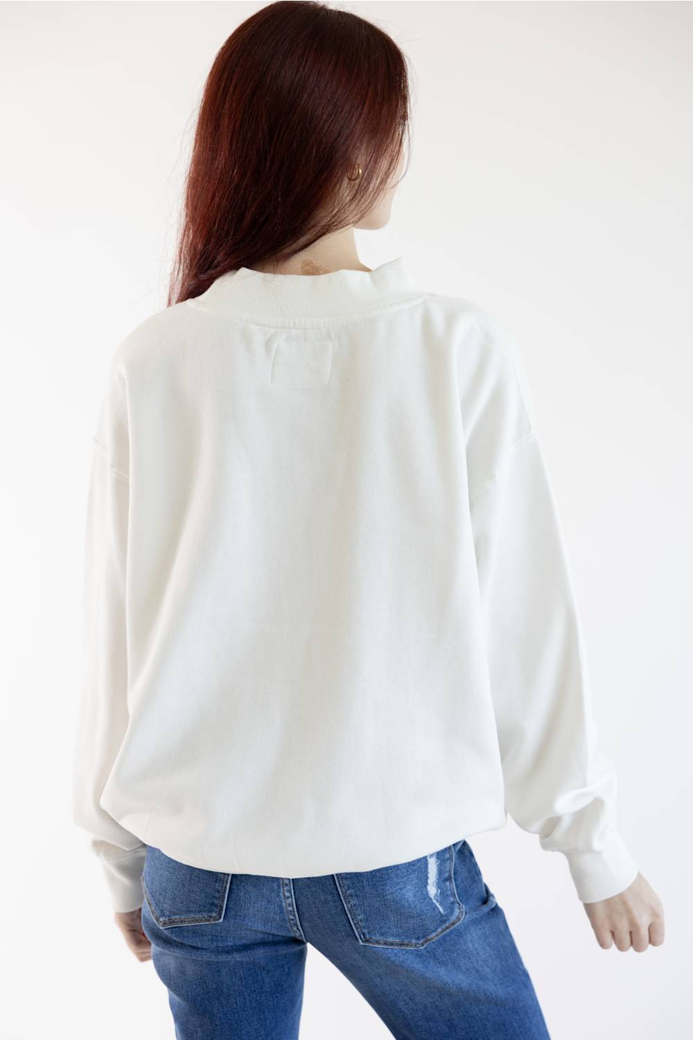 A woman viewed from behind wearing a loose white long-sleeve blouse and blue jeans against a plain white studio background.