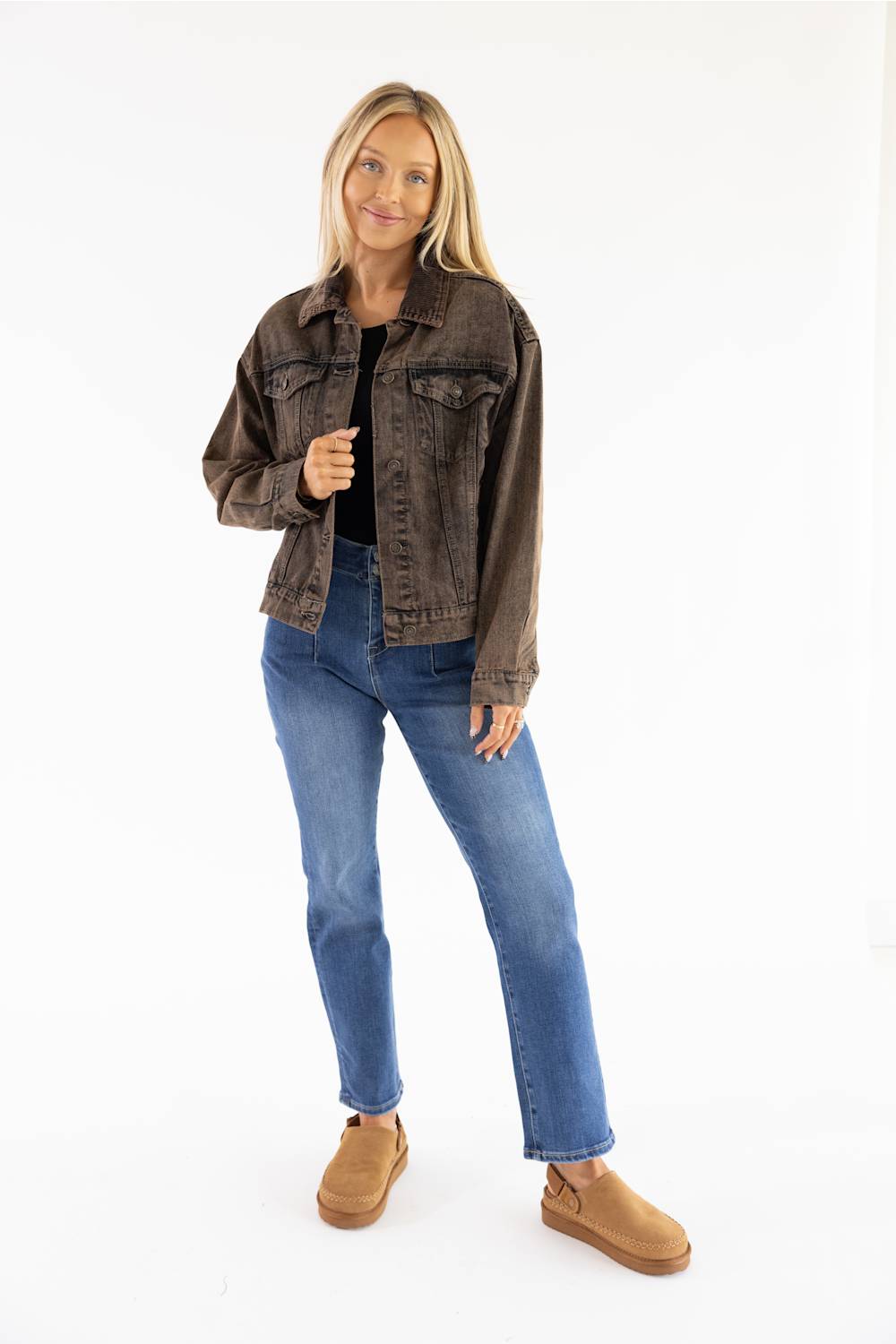A woman modeling a dark brown washed denim jacket, blue jeans, and tan slip-on boots against a plain white studio background.