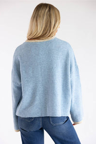 A woman viewed from behind wearing a light blue knit sweater with beige ribbed trim at the collar and cuffs, paired with blue jeans, against a plain white studio background.