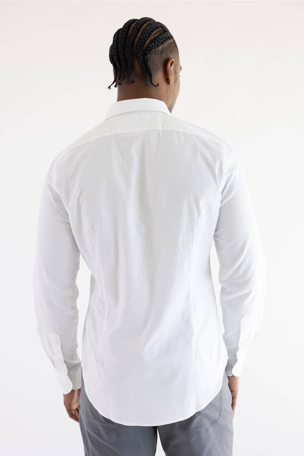 A man viewed from behind wearing a white long-sleeve button-down shirt and gray trousers, standing against a plain white studio background.