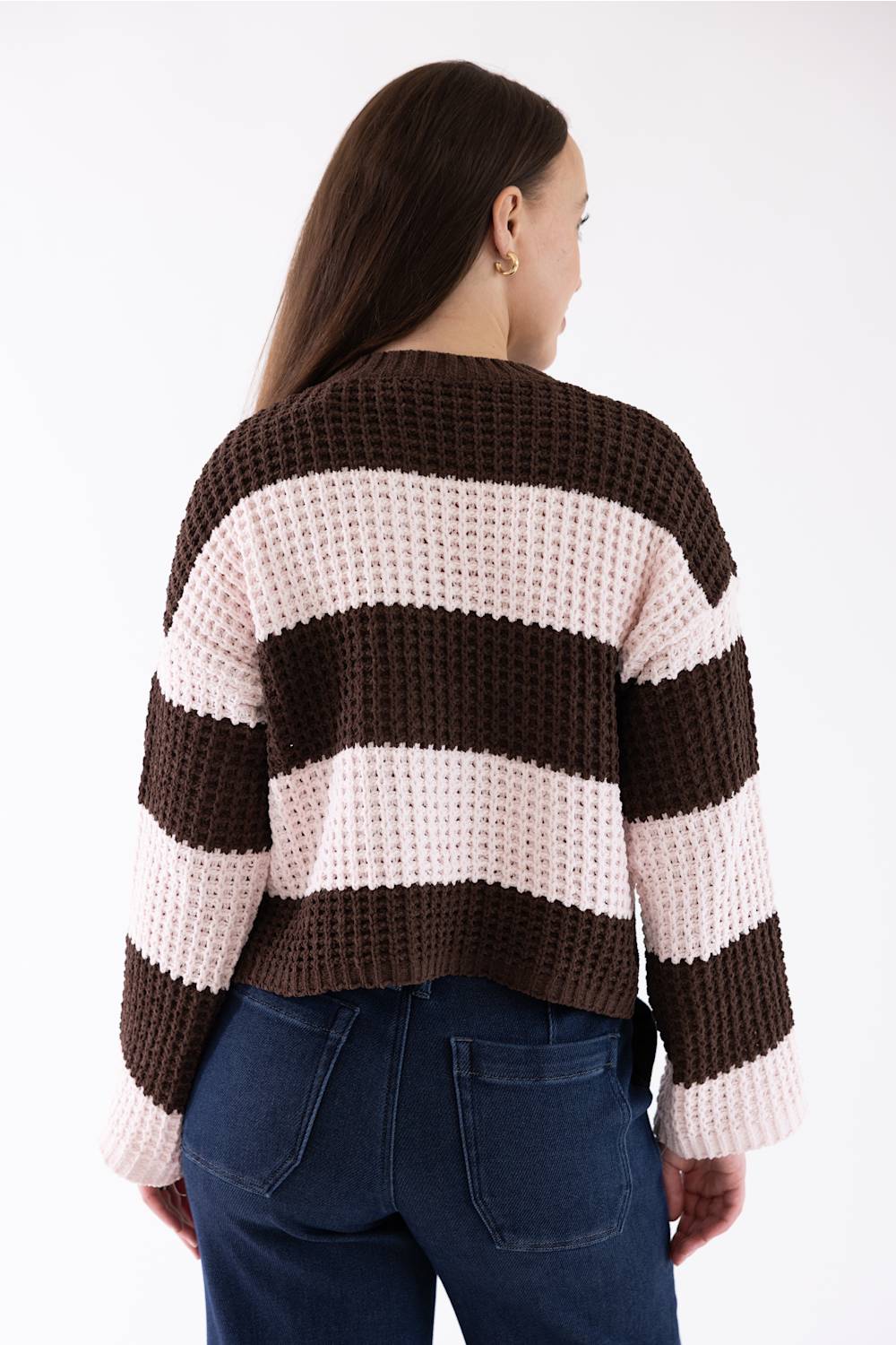 A woman from the back wearing a brown and light pink striped knit crew neck sweater and dark blue jeans against a white background.