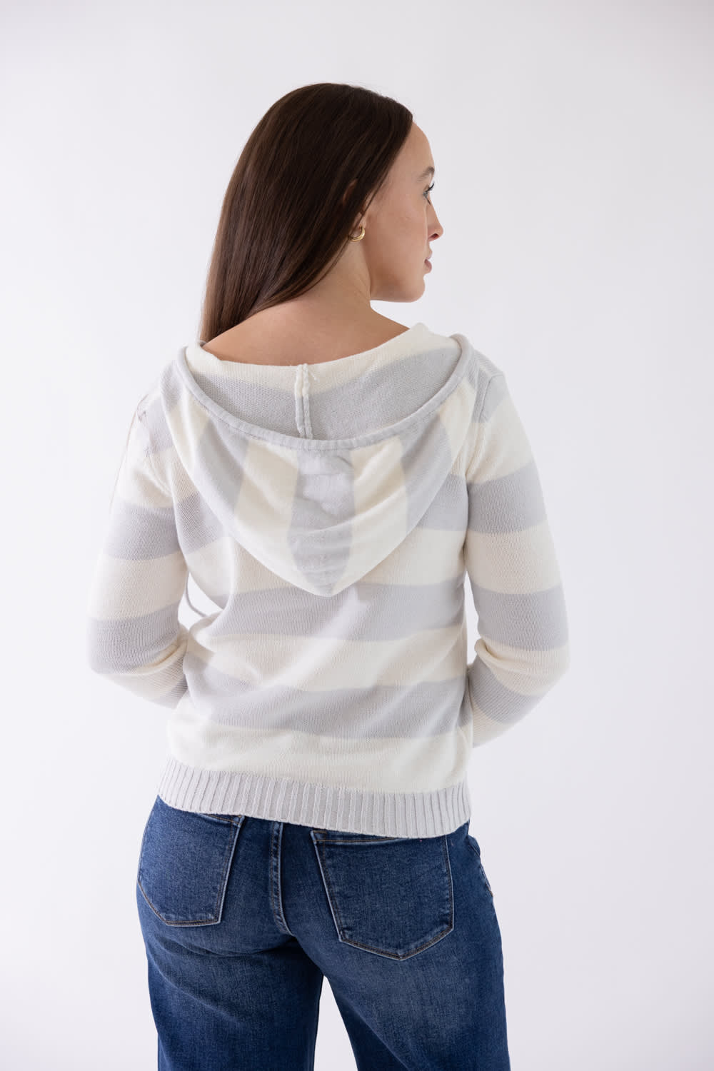 A woman shown from the back wearing a cream and light gray striped hooded sweater and blue jeans against a plain white studio background.