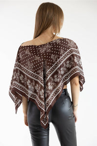 Back view of a model wearing a brown asymmetric scarf poncho top featuring a white floral and polka dot print, paired with black leather pants against a white background.