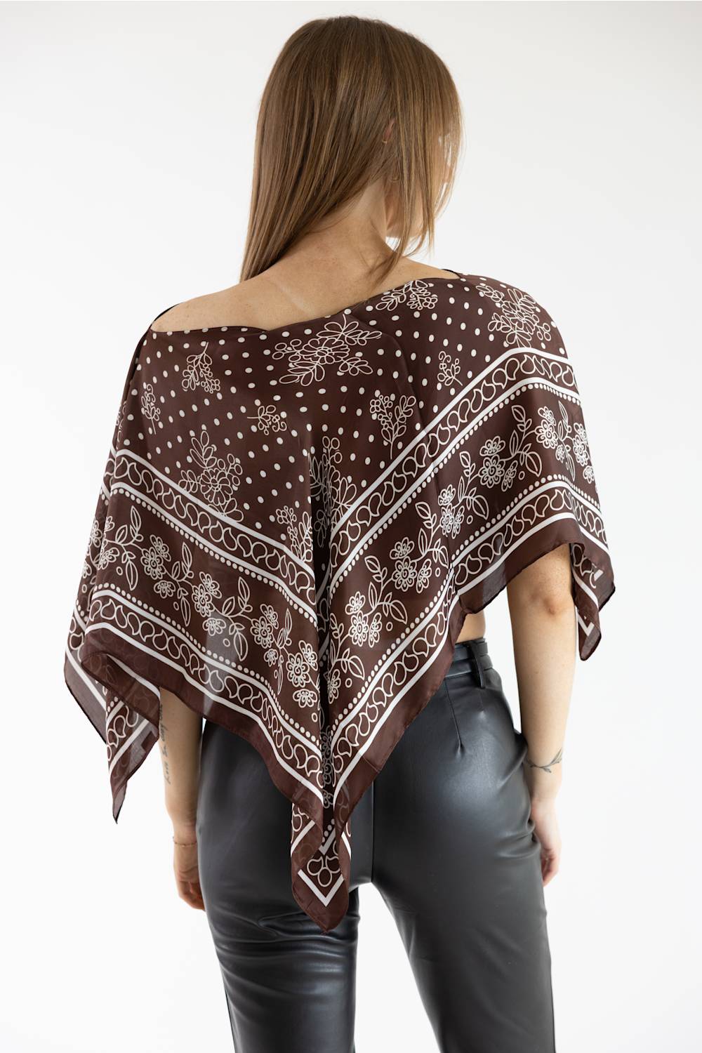 Back view of a model wearing a brown asymmetric scarf poncho top featuring a white floral and polka dot print, paired with black leather pants against a white background.