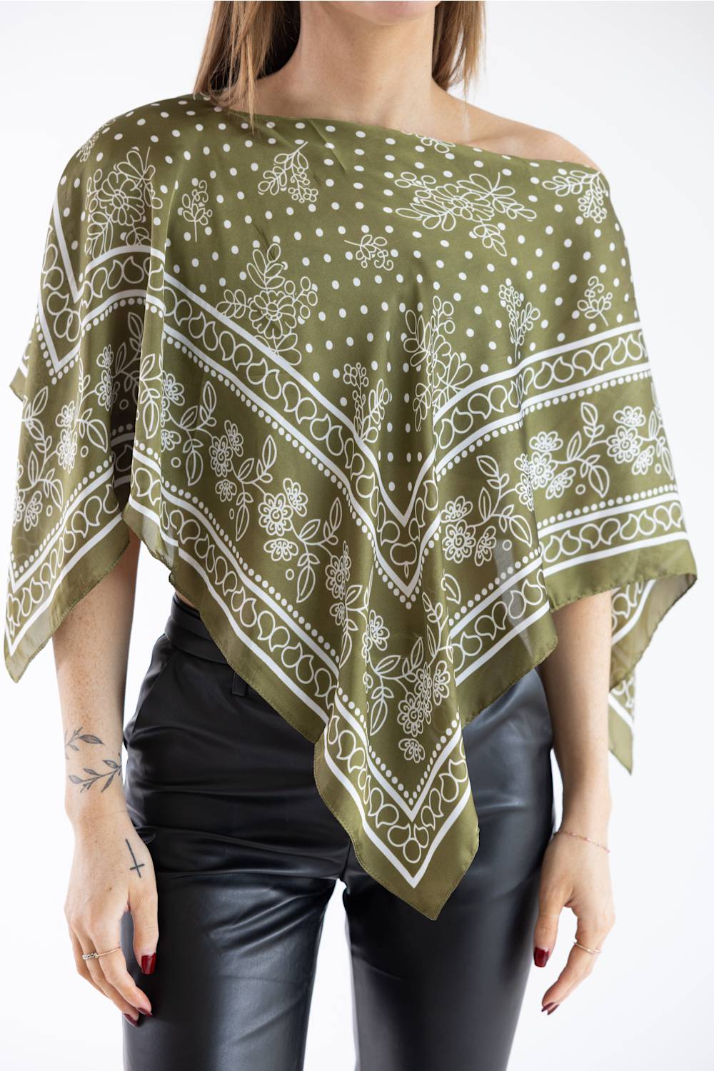 Close-up front view of a model wearing an olive green asymmetric scarf poncho top with a white floral and polka dot print, styled off one shoulder with black leather pants against a plain white studio background