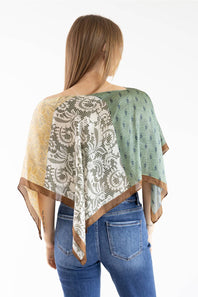 Back view of a model with straight brown hair wearing a patchwork scarf poncho top featuring yellow, white lace-patterned, and green floral panels with brown trim, styled with blue jeans against a white studio background.