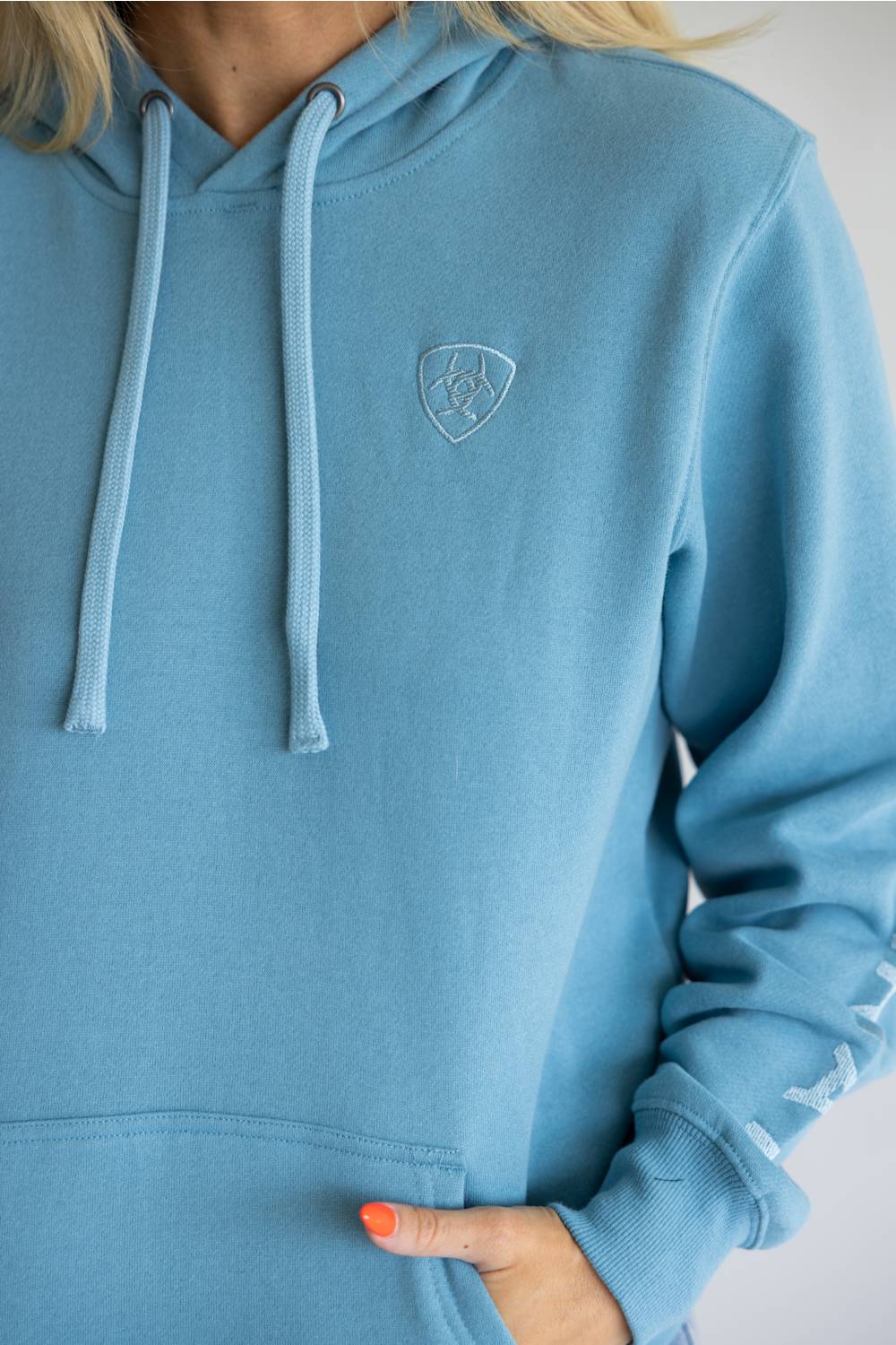 A close-up of a person wearing a light blue hooded sweatshirt. The hoodie has matching light blue drawstrings, a small shield-shaped embroidered logo on the left chest, and ribbed cuffs and hem.