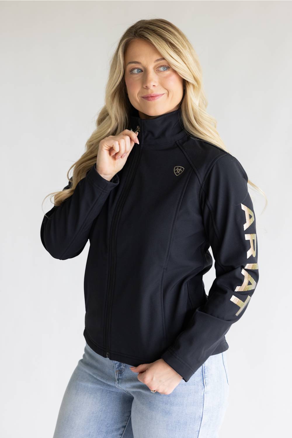 A woman wearing a black full-zip jacket with a high collar, featuring a small shield logo on the left chest and the brand name 'ARIAT' printed vertically in gold on the right sleeve, paired with light blue jeans against a light gray background.