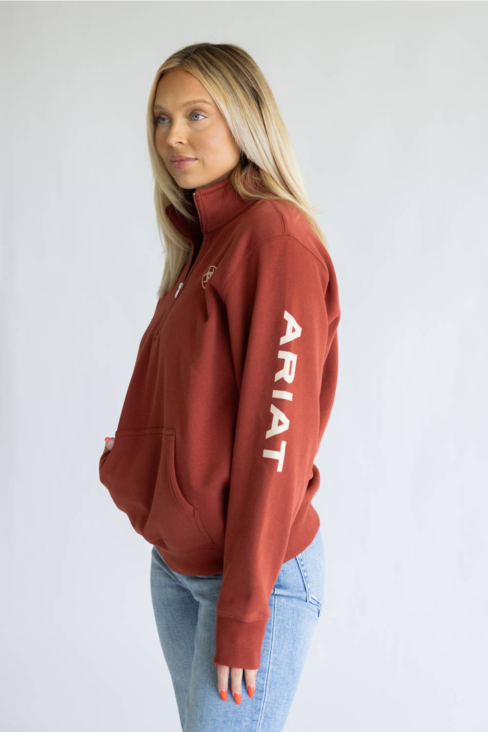 A woman modeling a rust-colored quarter-zip hoodie with 'ARIAT' printed vertically in white on the sleeve, paired with light blue jeans against a plain light gray background.