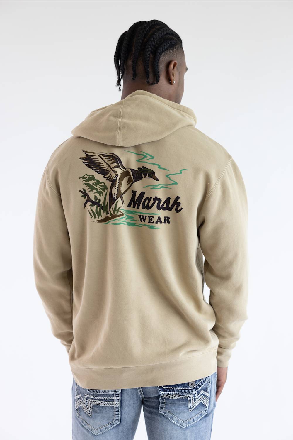 A man viewed from behind wearing a beige hoodie with a large graphic on the back featuring a duck, plants, and the text 'Marsh Wear', paired with blue jeans against a light gray studio background.